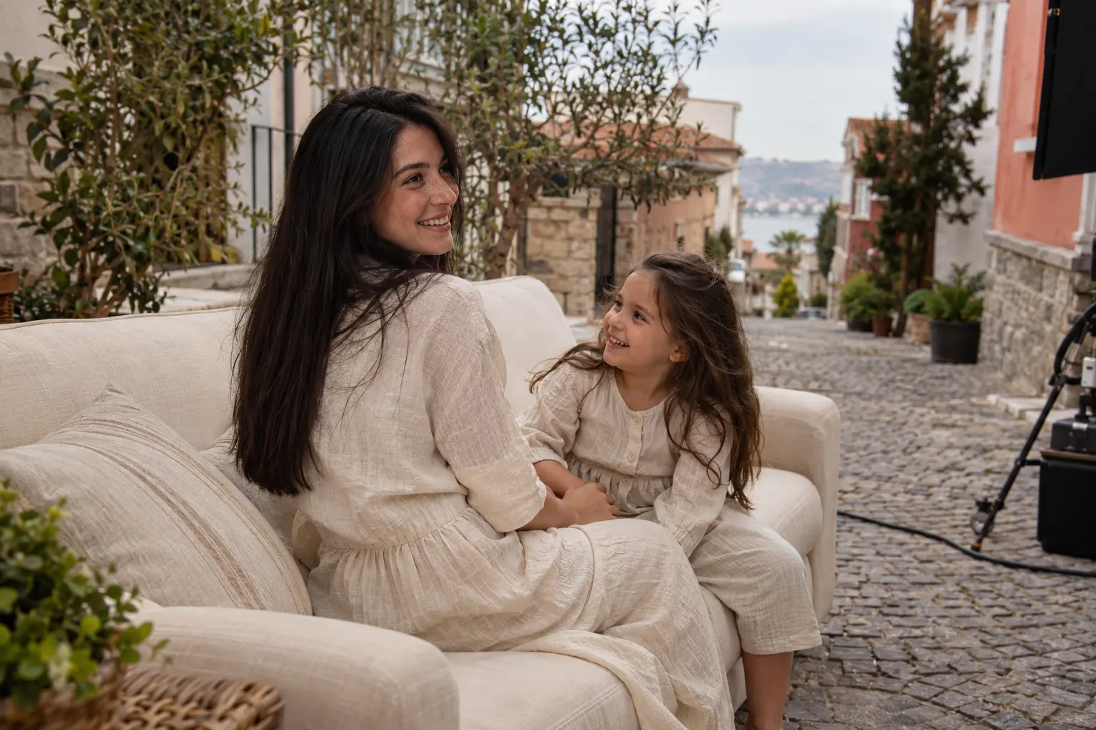 Anne ve küçük kızı, taş sokaktaki katalog çekiminde krem koltukta birlikte gülüyor.