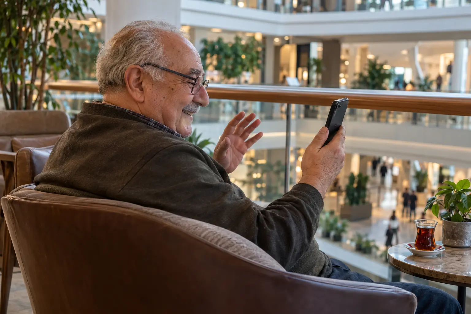 AVM balkonunda oturan gözlüklü yaşlı adam, görüntülü görüşme sırasında telefonuna gülümsüyor.